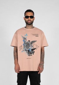 Lys pink oversized t-shirt med en grafisk tryk af en engel og en figur, med blå accenter og tekst på forsiden. Korte ærmer, blød stof.