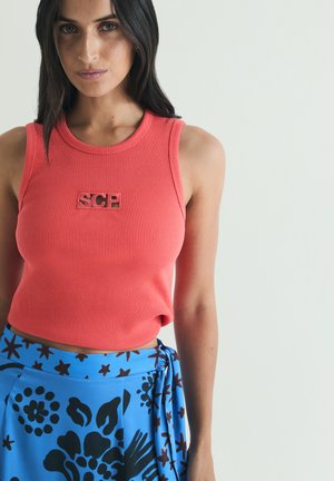 Femme portant un haut côtelé corail sans manches avec l'inscription "SCP" et une jupe portefeuille bleue à motifs floraux et étoiles noirs.