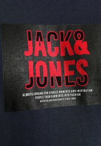 Námorná tkanina s čiernym obdĺžnikovým štítkom. Na ňom je výrazný červený a biely text "JACK & JONES" spolu s podtitulom nižšie.