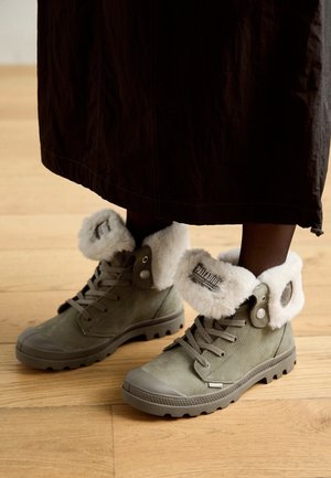 Oliijfgroene veter-winterlaarzen met witte bontvoering, gedragen met donkere kleding op een houten vloer.