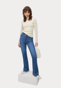 Top lungo a maniche, in crema chiaro e texturizzato, con collo alto; jeans a zampa d'elefante blu; scarpe argento a punta; tiene una borsa a rete di forma arrotondata.