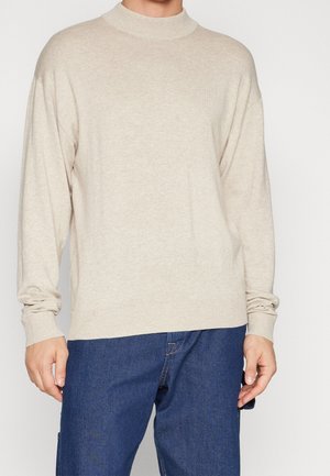 Suéter beige de manga larga llevado con jeans de mezclilla azules en una persona de pie, cortado a la altura del cuello y a mitad del muslo.