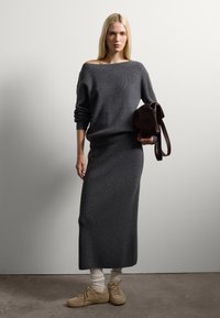 Suéter gris de punto con un diseño de hombros descubiertos, combinado con una falda larga a juego. Un bolso marrón y zapatillas de color claro completan el look.