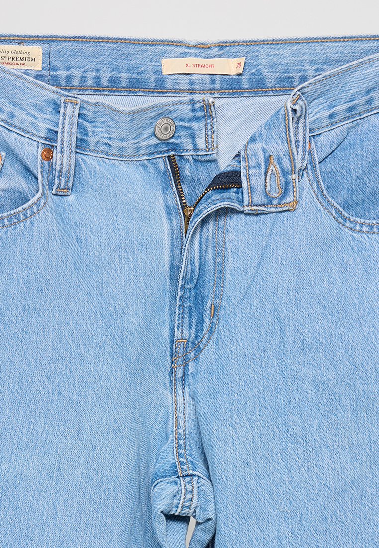 Jeans en denim bleu clair avec une coupe droite. Comprend une fermeture éclair, un bouton de fermeture et des détails de surpiqûres orange. L'étiquette indique "XL DROIT".
