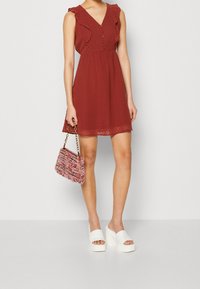 Kobieta ubrana w bezrękawową sukienkę w kolorze rdzy z guzikami i falbanami, trzymająca różową torebkę o fakturze oraz białe sandały na platformie.