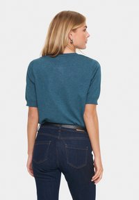 Mėlynos-žalios spalvos trumpas rankovėmis megztinis su apvaliu iškirpimu ir raukšlėtą tekstūrą, derinamas su tamsiai mėlynomis džinsomis ir juodu diržu.