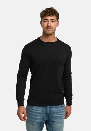 Junger Mann mit kurzen lockigen Haaren, der einen schwarzen Pullover mit langen Ärmeln und blaue Jeans trägt und vor einem weißen Hintergrund steht.