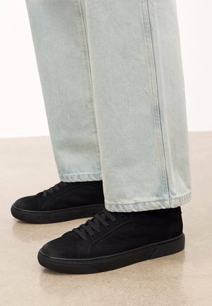 Zapatillas de ante negro con punta redonda, parte superior texturizada y suela de goma plana, combinadas con pantalones de mezclilla azul claro.