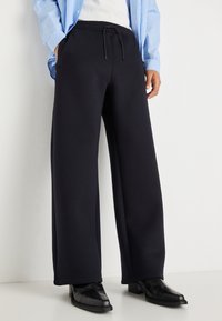 Marineblaue, weit geschnittene Sweatpants aus weichem Material, mit einem elastischen Bund und Kordelzug sowie seitlichen Taschen, kombiniert mit schwarzen spitzen Schuhen.
