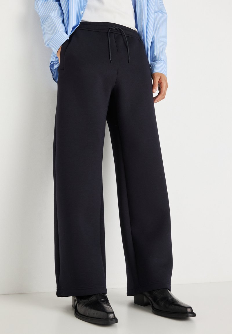 Marineblaue, weit geschnittene Sweatpants aus weichem Material, mit einem elastischen Bund und Kordelzug sowie seitlichen Taschen, kombiniert mit schwarzen spitzen Schuhen.