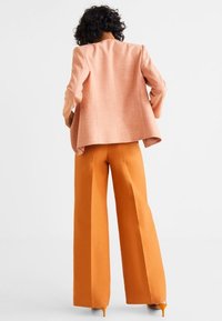 Person with curly hair wearing a peach blazer and wide-legged orange pants, standing with hands on hips against a plain background.