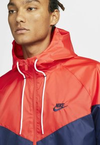 Nike vėjara su ryškiai raudonu viršumi ir tamsiai mėlynomis apatinėmis dalimis, su užtrauktuku priekyje, reguliuojamu raišteliu ant kapučino ir logotipo detale.