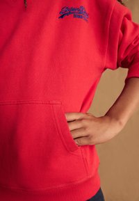 Červená mikina s texturovaným povrchem, má klokánkovou kapsu a modrou vyšívanou značku "SuperDry" na hrudi. Rukávy jsou pružné.