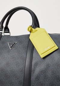 Sac à main gris texturé avec des accents en cuir noir, arborant un logo triangulaire et une étiquette de bagages jaune. Fermeture éclair sur le dessus.