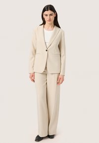 Completo beige con giacca aderente e pantaloni a gamba larga. La giacca presenta un bottone singolo e due tasche frontali. Tessuto dalla texture liscia.