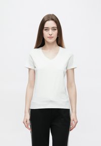 Άσπρο t-shirt με λαιμόκοψη σε σχήμα V, κατασκευασμένο από απαλό βαμβακερό ύφασμα, με κοντά μανίκια και άνετη εφαρμογή, συνδυασμένο με μαύρο παντελόνι. Απλός και ευέλικτος σχεδιασμός.