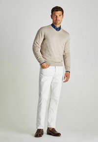 Beiger V-Ausschnitt-Pullover über einem blauen Hemd mit Kragen, kombiniert mit weißen Hosen und braunen Slippern. Glatte Texturen, moderne Passform, lässiger Look.