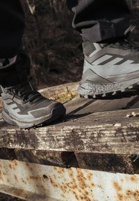 Stivali outdoor neri e grigi con materiale testurizzato, dotati di una suola robusta, lacci prominenti e dettagli a strisce diagonali, poggiati su legno stagionato.