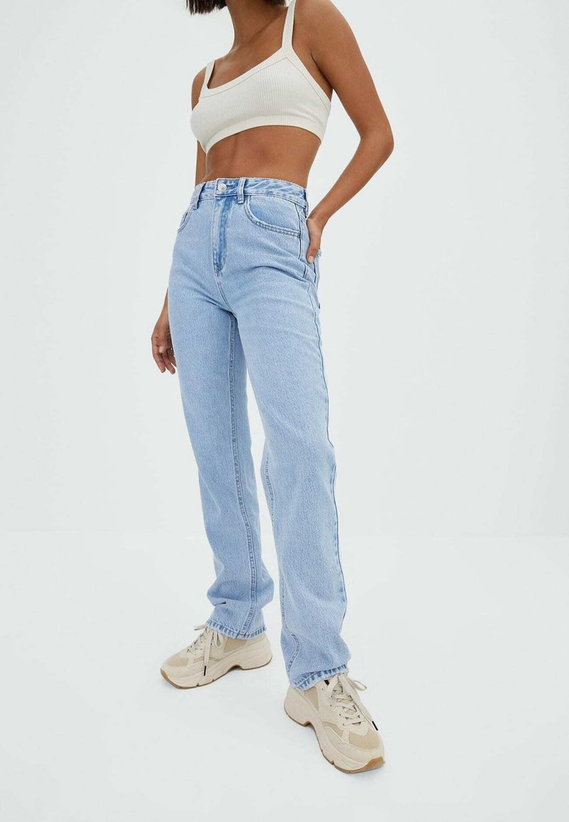 Personne portant un jean taille haute bleu clair, un crop top blanc et des baskets beiges, debout devant un fond blanc uni.