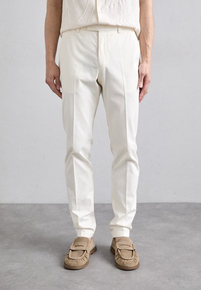 White tailored trousers made of smooth fabric, featuring a classic fit with front pleats, paired with beige suede loafers.
