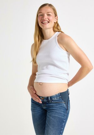 Vitt ribbat linne och blå denimjeans med ett stretchpanel i midjan, som visar en synlig gravidmage med händer som vilar på den.