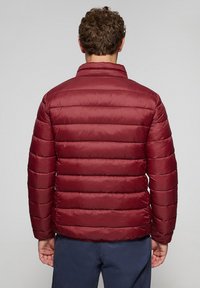 Chaqueta acolchada color burdeos con costuras horizontales, cuello alto y material liviano. Presenta una textura suave y un diseño ajustado.