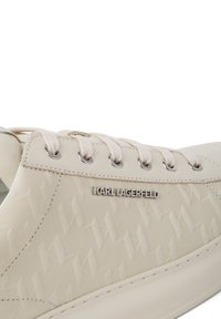 Beežid tossud tekstureeritud pealsega, millel on korduv logomuster, kontrastsed heledad paelad ja hõbedane brändiplaat küljes.