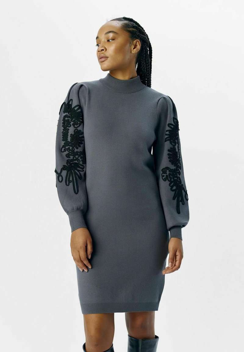 Robe en tricot gris à manches longues et bouffantes, ornée de broderies florales noires. La robe a un col montant et des poignets côtelés, offrant une silhouette ajustée.