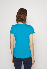 Kvinna med kort brunt hår, klädd i en åtsittande turkos kortärmad skjorta och mörkblå byxor, som står med ryggen mot en enkel bakgrund.