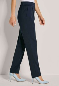 Vrouw in marineblauwe op maat gemaakte broek en lichtblauwe hoge hakken, staande tegen een effen lichte achtergrond.