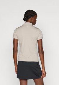Lys beige polo shirt med krave, korte ærmer og et gyldent logo på ryggen. Båret med en sort nederdel, der fremviser en glat tekstur.
