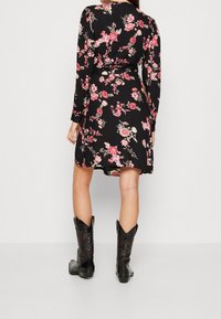 Kvinna i en svart blommig klänning med lång ärm, kombinerad med svarta broderade cowboystövlar, stående mot en enkel ljus bakgrund.