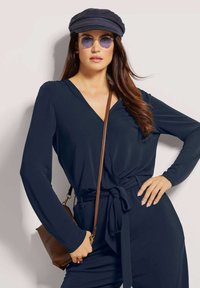 Donkerblauwe langlopende wikkeltop met een V-hals en geplooide details bij de buste, gecombineerd met een bruine crossbody tas en accessoires.