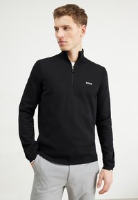 Black zip-up sweater with ribbed accents and a small white logo on the chest, paired with light gray trousers. Smooth fabric texture.