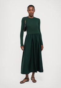 Long-sleeved, dark green dress made of knit fabric, featuring a fitted top and a gathered, flowing skirt. Worn with black handbag and sandals.