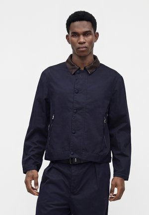 Jeune homme portant une veste boutonnée bleu marine foncé avec un col marron et un pantalon bleu marine assorti sur un fond clair uni.