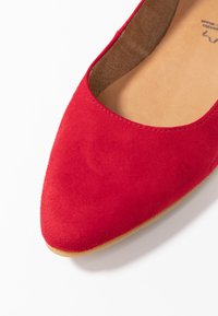 Chaussure plate en suède rouge avec un bout pointu, présentant une texture lisse et un intérieur doux. La semelle est plate et légèrement rembourrée.