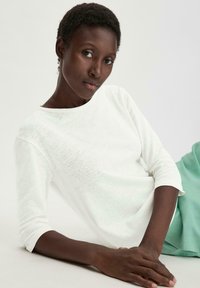 Weißes, strukturiertes Langarmoberteil mit einem Boot-Ausschnitt, kombiniert mit einem mintgrünen Rock, getragen von einem Model mit kurzen Haaren.