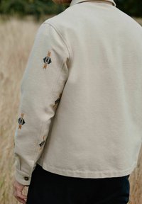 Beige katoenen jas met een kraag en knoopmanchetten, met zwarte en oranje geometrische borduursels op de mouwen.