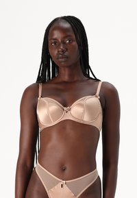 Γυμνός σατέν σουτιέν με βραχίονα και λεπτομέρεια φιόγκου στο κέντρο, που διαθέτει ρυθμιζόμενους ιμάντες και λεία υφή. Συμπεριλαμβάνεται αντίστοιχο διάφανο στρινγκ.