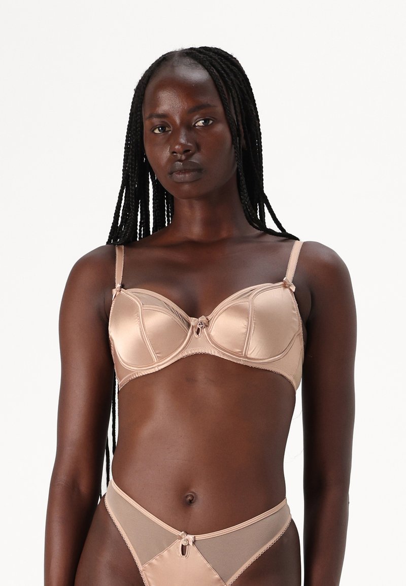 Γυμνός σατέν σουτιέν με βραχίονα και λεπτομέρεια φιόγκου στο κέντρο, που διαθέτει ρυθμιζόμενους ιμάντες και λεία υφή. Συμπεριλαμβάνεται αντίστοιχο διάφανο στρινγκ.