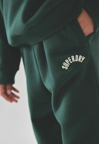 Felpa e pantaloni verdi in tessuto morbido, con una texture a coste. Dettaglio notevole: logo "SUPERDRY" ricamato in colore crema.
