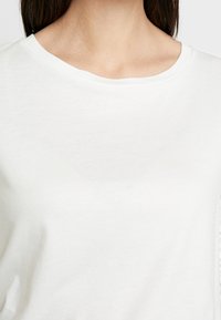 Weißes, kurzärmliges Oberteil aus weichem Baumwollmaterial. Verfügt über einen runden Ausschnitt und eine glatte Textur ohne sichtbare Muster oder Verzierungen.