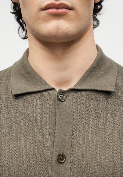 Το κάτω μέρος του προσώπου και ο λαιμός ενός άνδρα που φοράει καφέ πλεκτό πόλο με δύο κουμπιά κλειστά.