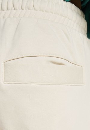 Light cream-colored joggers featuring a textured waistband and a square back pocket with a stitched border. Soft cotton blend material.