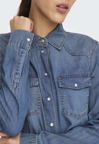 Camisa de mezclilla en azul claro, con un frente de botones, dos bolsillos en el pecho y costuras en contraste. Con cuello y botones plateados.