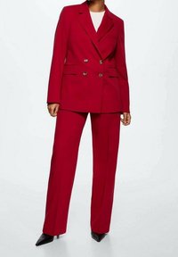 Personne portant un costume rouge à double boutonnage avec un pantalon à jambes larges et des escarpins noirs à bout pointu, se tenant devant un fond blanc uni.