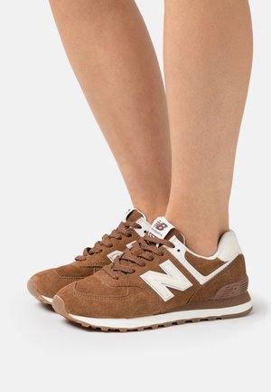 Baskets New Balance en daim marron avec logo et accents blancs, portées pieds nus sur un fond blanc.