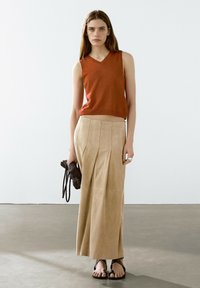 Top senza maniche color ruggine, gonna maxi plissettata in suede beige, tiene una borsa nera texturizzata e indossa sandali neri piatti.