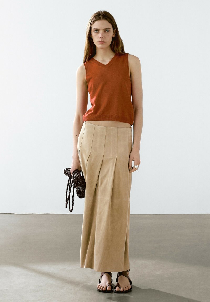 Top senza maniche color ruggine, gonna maxi plissettata in suede beige, tiene una borsa nera texturizzata e indossa sandali neri piatti.
